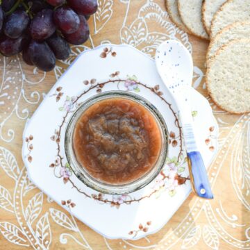 A jar of rhubarb chutney on a wooden tray with grapes and oatcakes.